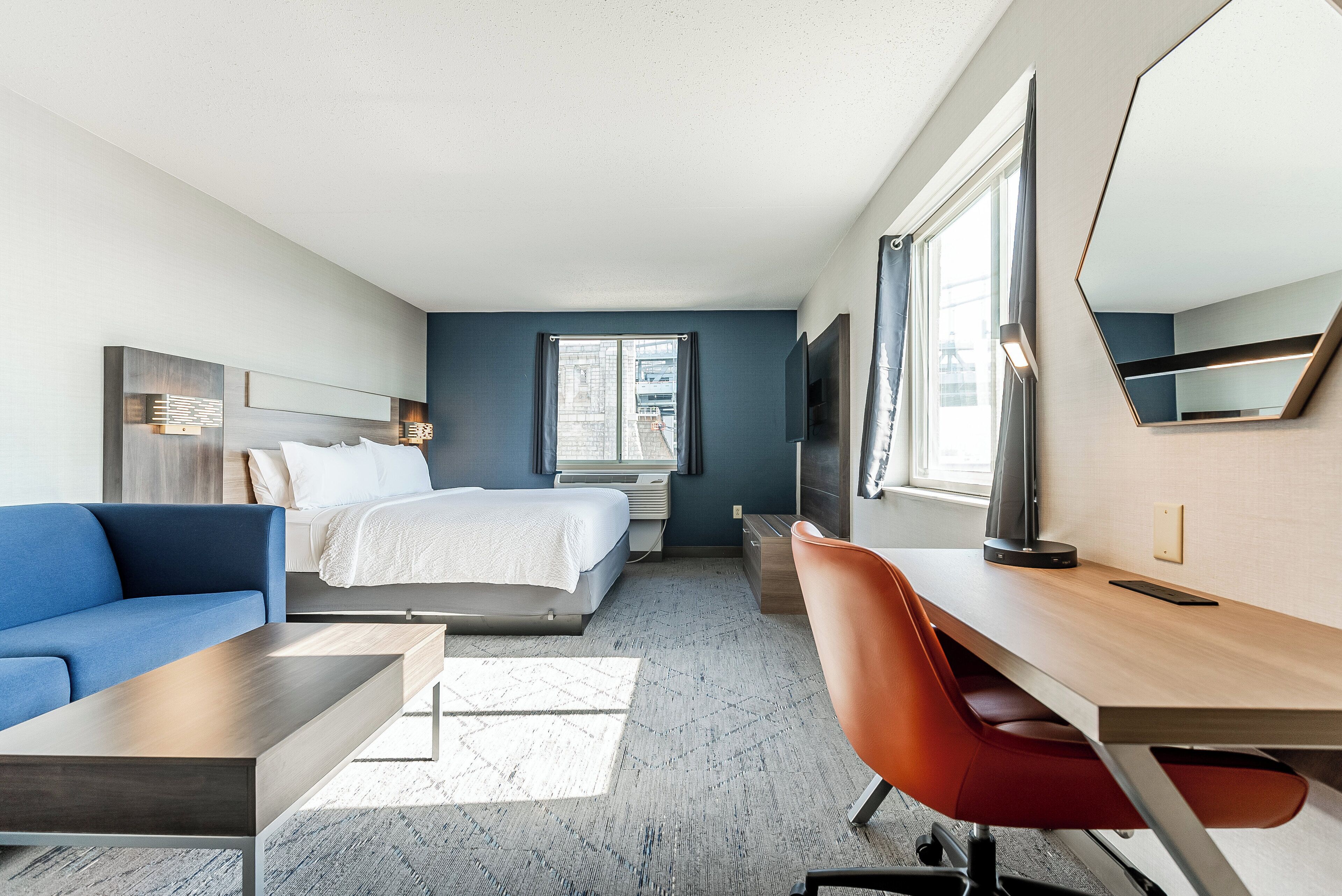 Una habitación de hotel moderna con una cama, un sofá azul, un escritorio de madera y un espejo grande.