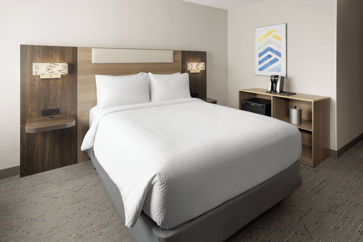 Quarto de hotel moderno com uma cama grande, cabeceira de madeira, mesas de cabeceira, um criado-mudo e uma máquina de café.