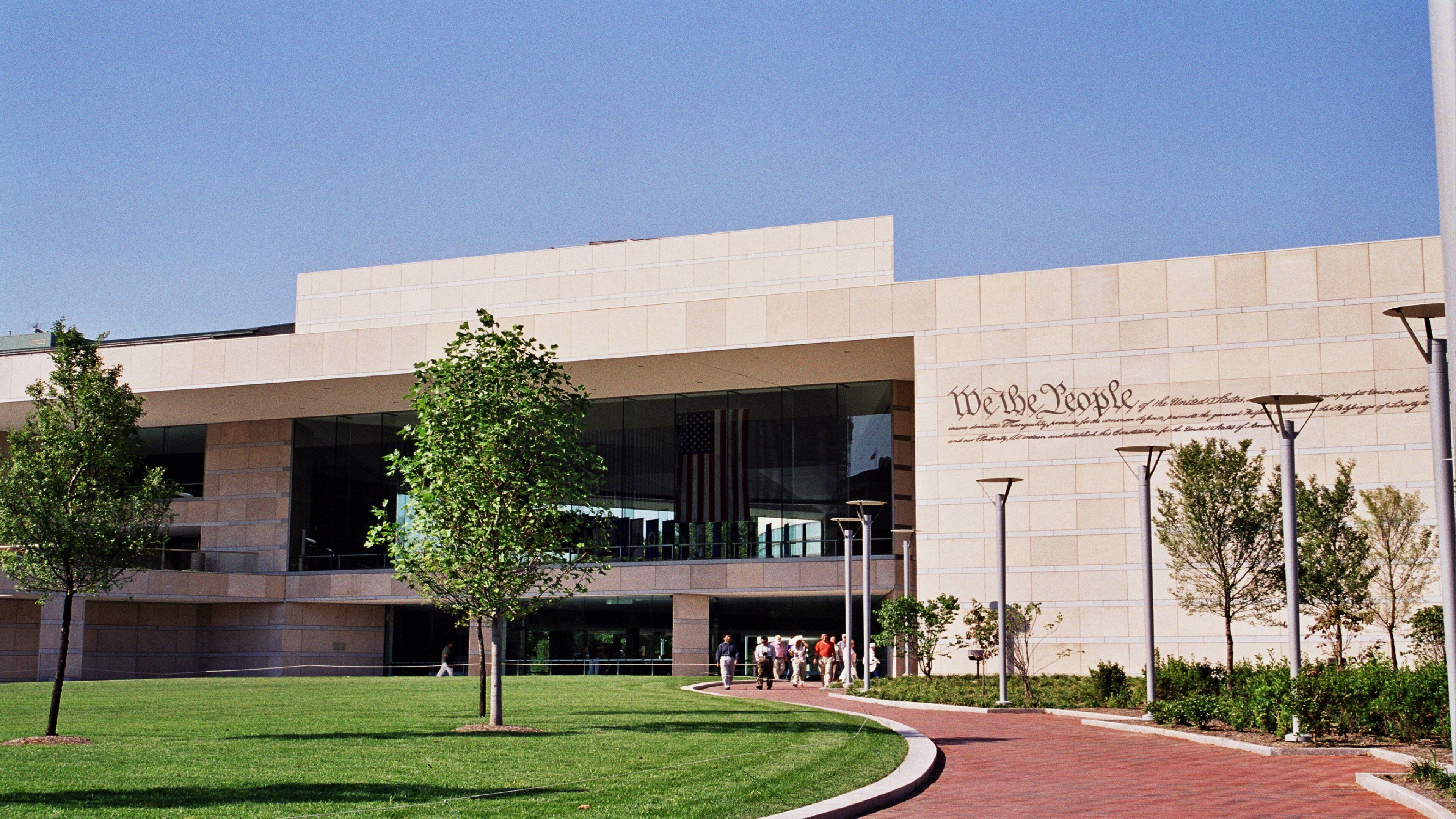 A modern building with a large glass facade, a landscaped area with trees, and a paved walkway.