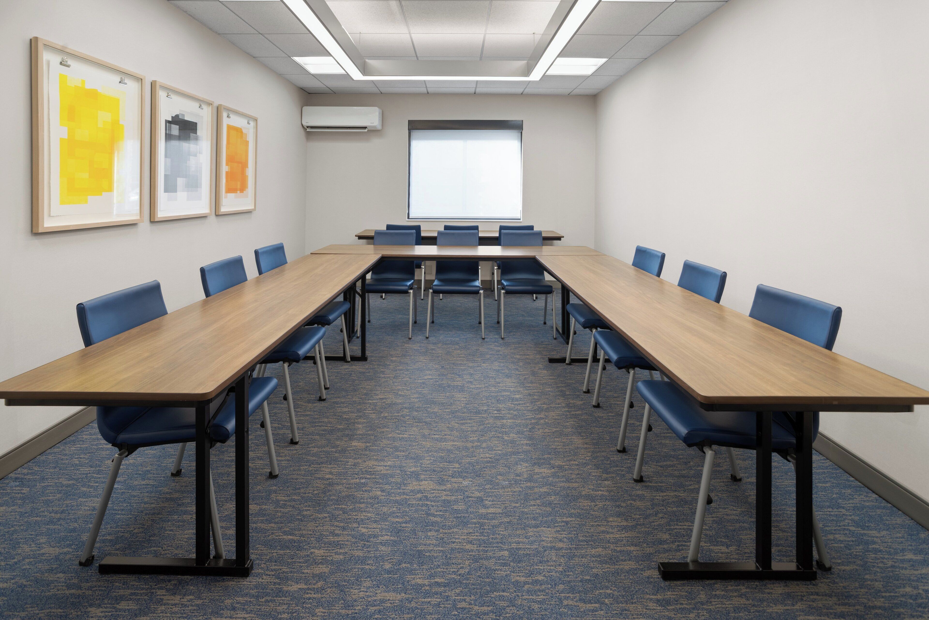 A conference room with a long table, blue chairs, framed artwork on the wall, and a large window.