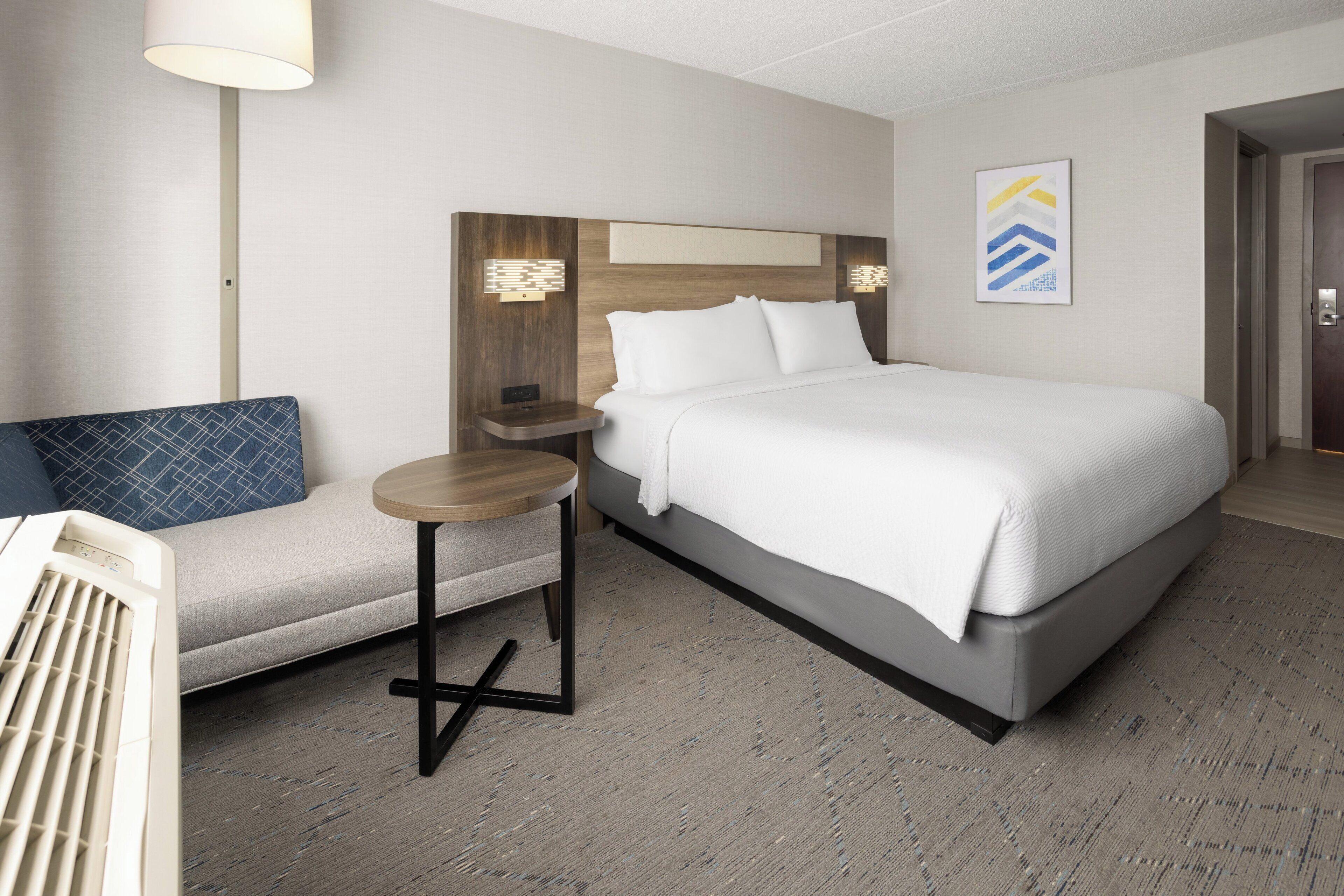 A hotel room with a large bed, a small table, a sofa, and a wall-mounted lamp.