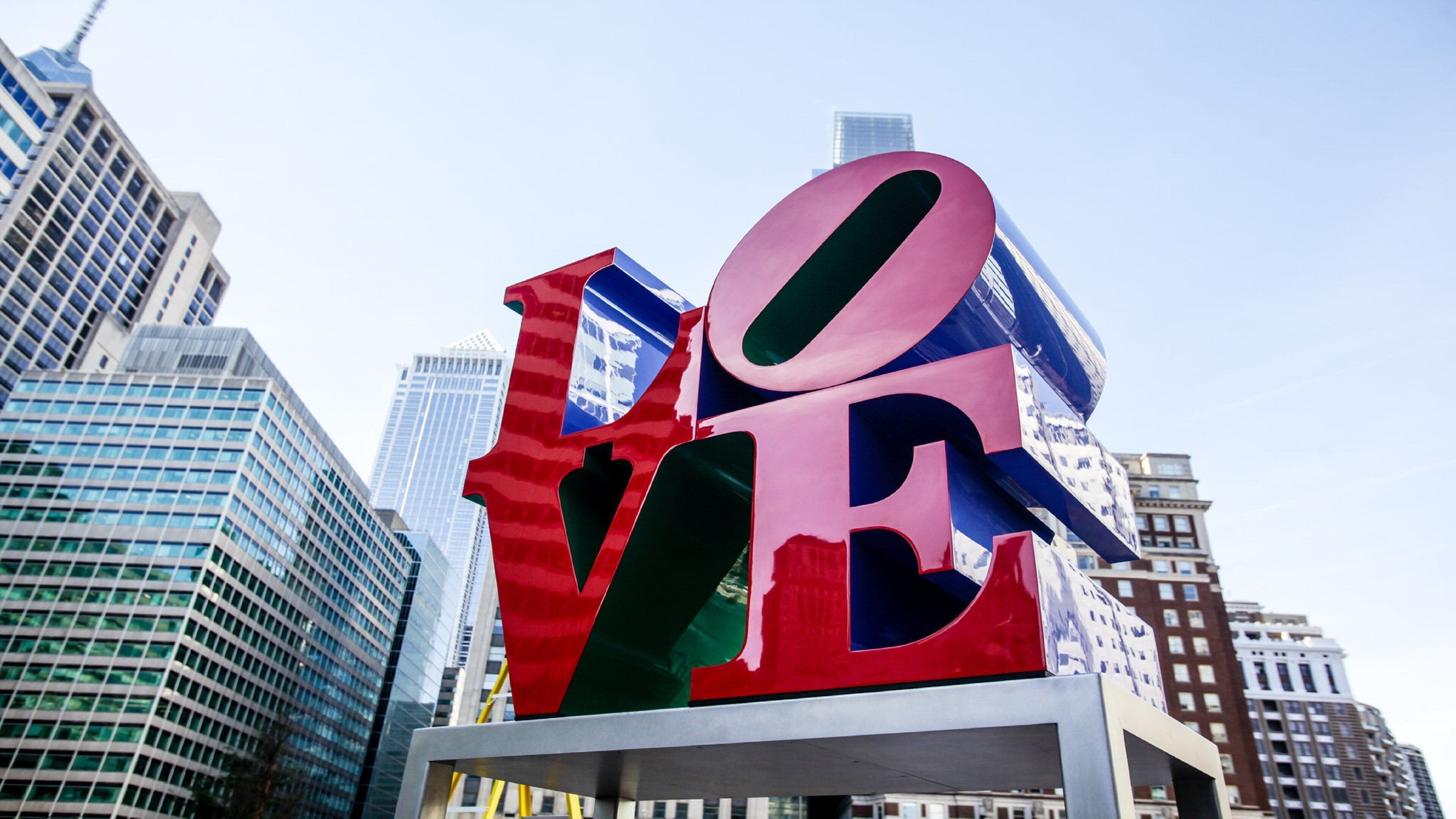 A large, colorful sculpture with the letters 'LOVE' in the foreground, set against a backdrop of modern skyscrapers.