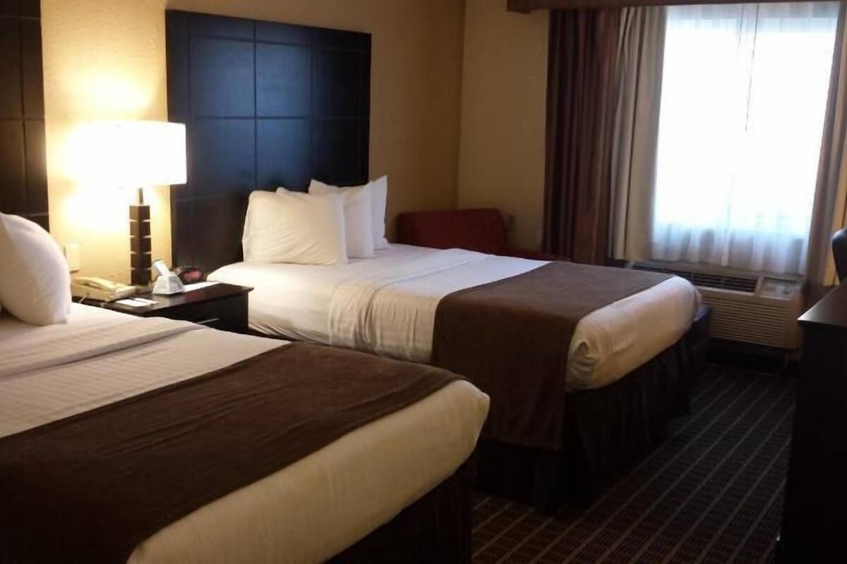 A hotel room with two beds, a nightstand with a lamp, a desk, and a window with curtains.
