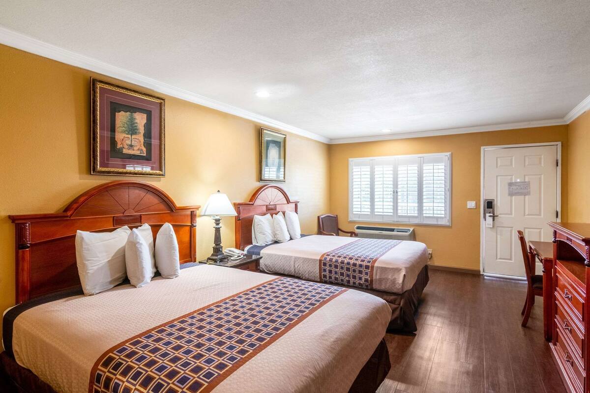 Une chambre d’hôtel avec deux lits, un bureau, une chaise, une lampe et une fenêtre avec des stores.