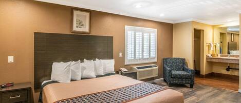 Habitación de hotel con cama, una silla, un escritorio, una ventana con persianas, un cuadro en la pared y un baño visible a través de una puerta abierta.