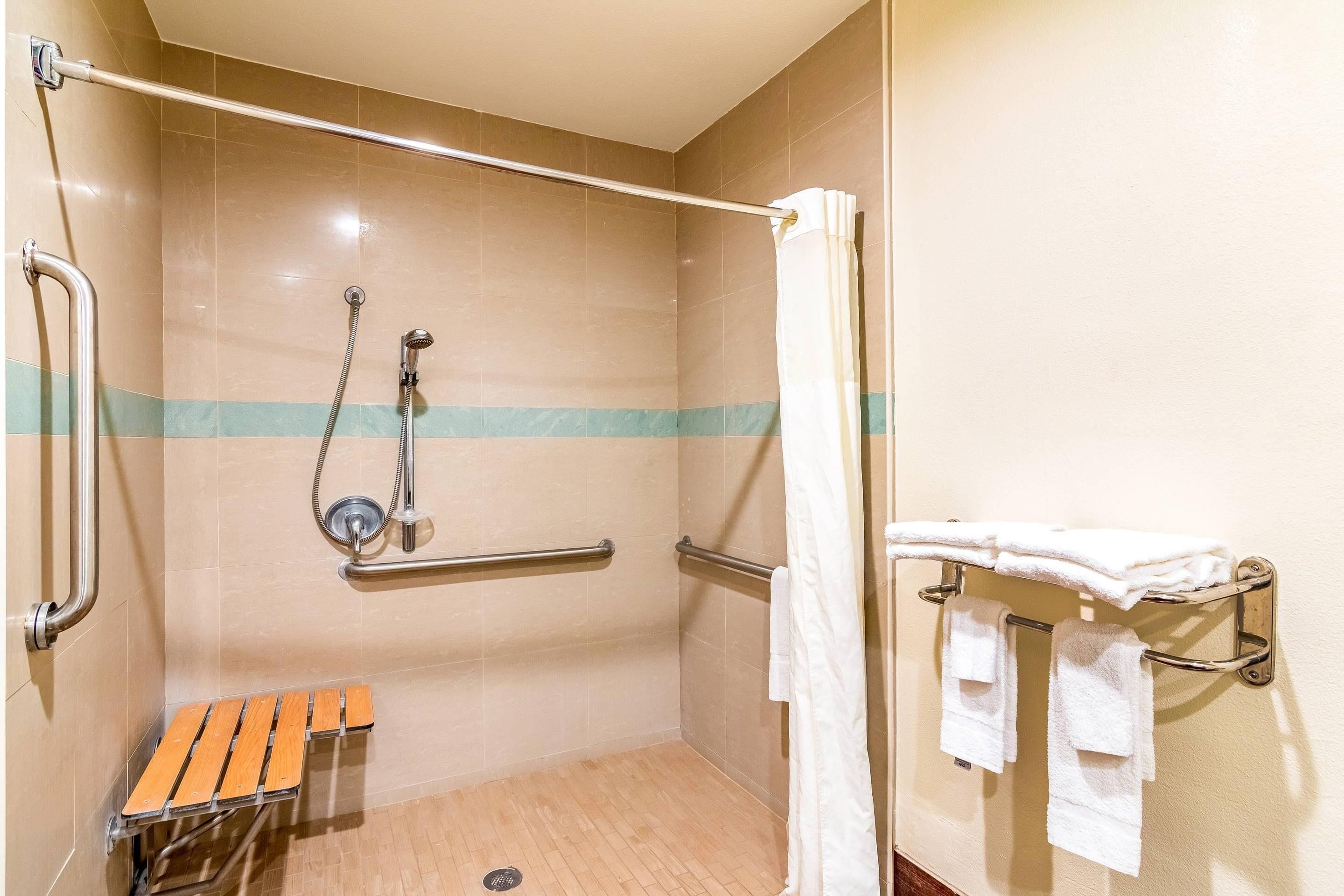 A shower area with a handrail, a wooden bench, a towel rack with towels, and a showerhead.