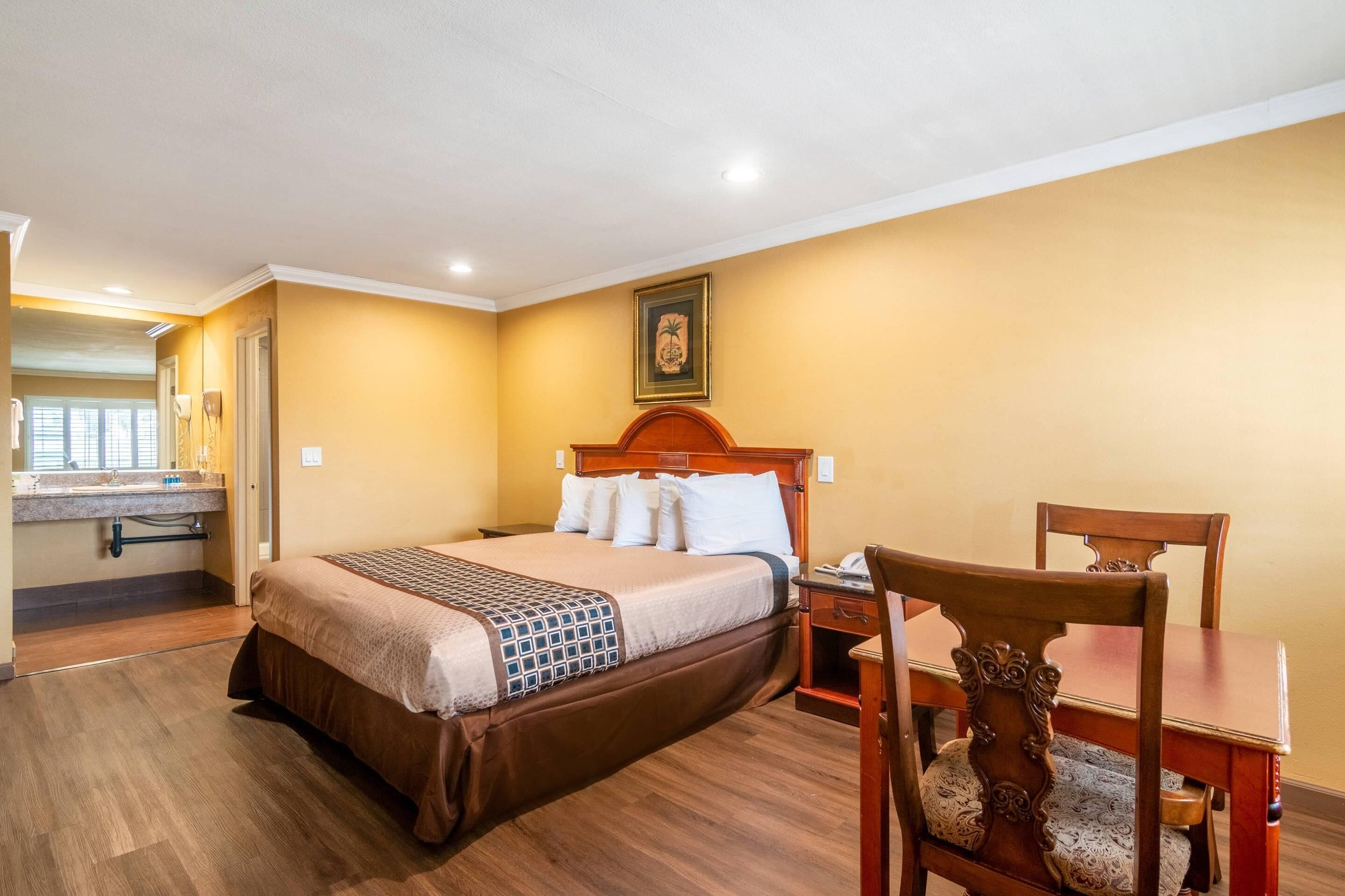 A hotel room with a bed, a wooden chair, a small table, a nightstand, and a bathroom visible through an open door.