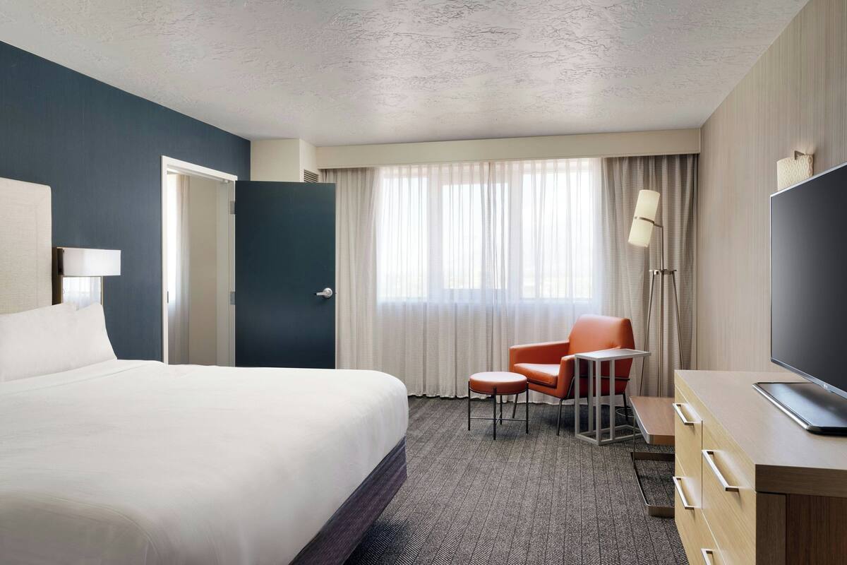 A hotel room with a large bed, a desk with a flat-screen TV, a chair, and a desk lamp.