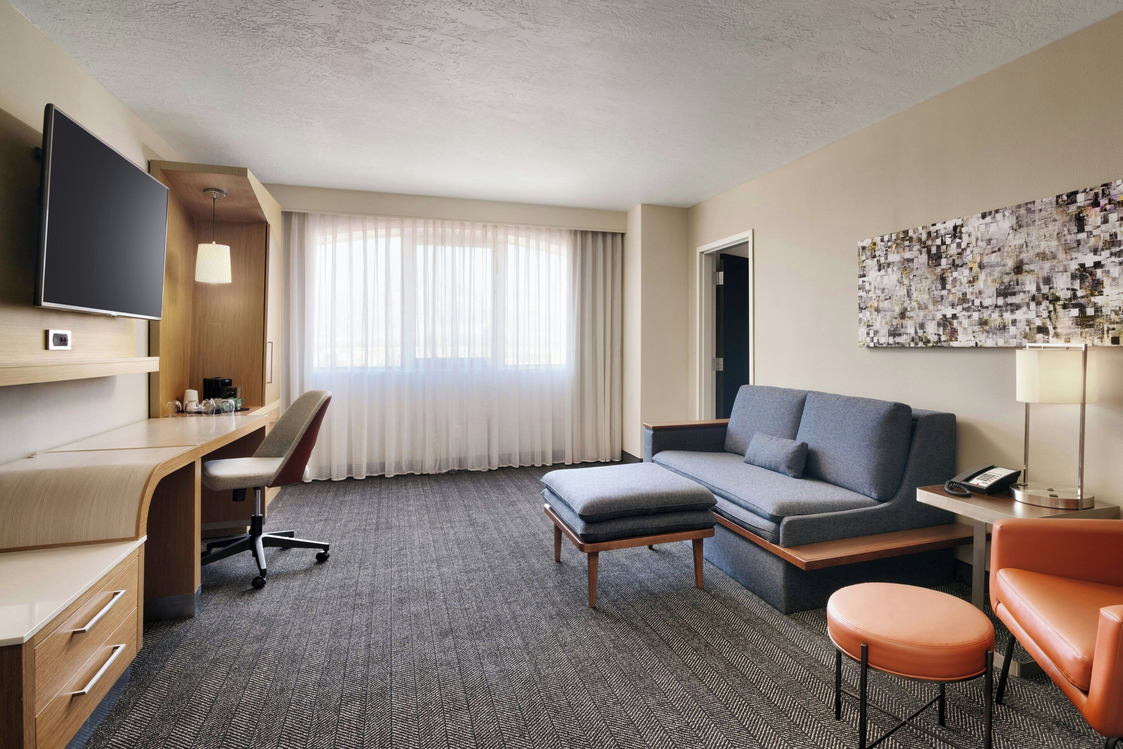 Habitación de hotel con escritorio, sofá, otoman, televisor y una ventana grande con cortinas.