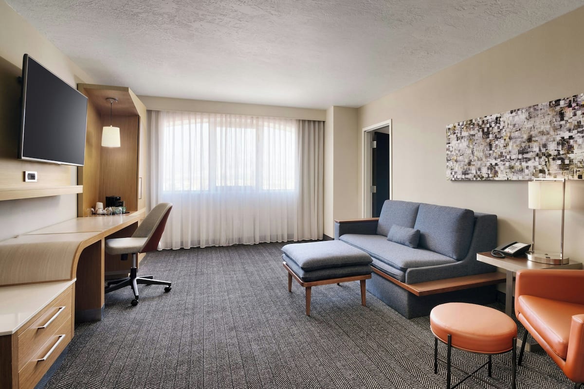 A hotel room with a desk, a sofa, an ottoman, a TV, and a large window with curtains.