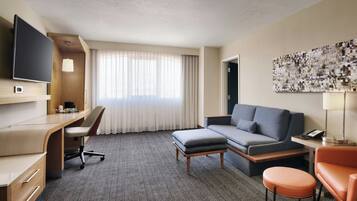 Habitación de hotel con escritorio, sofá, otoman, televisor y una ventana grande con cortinas.