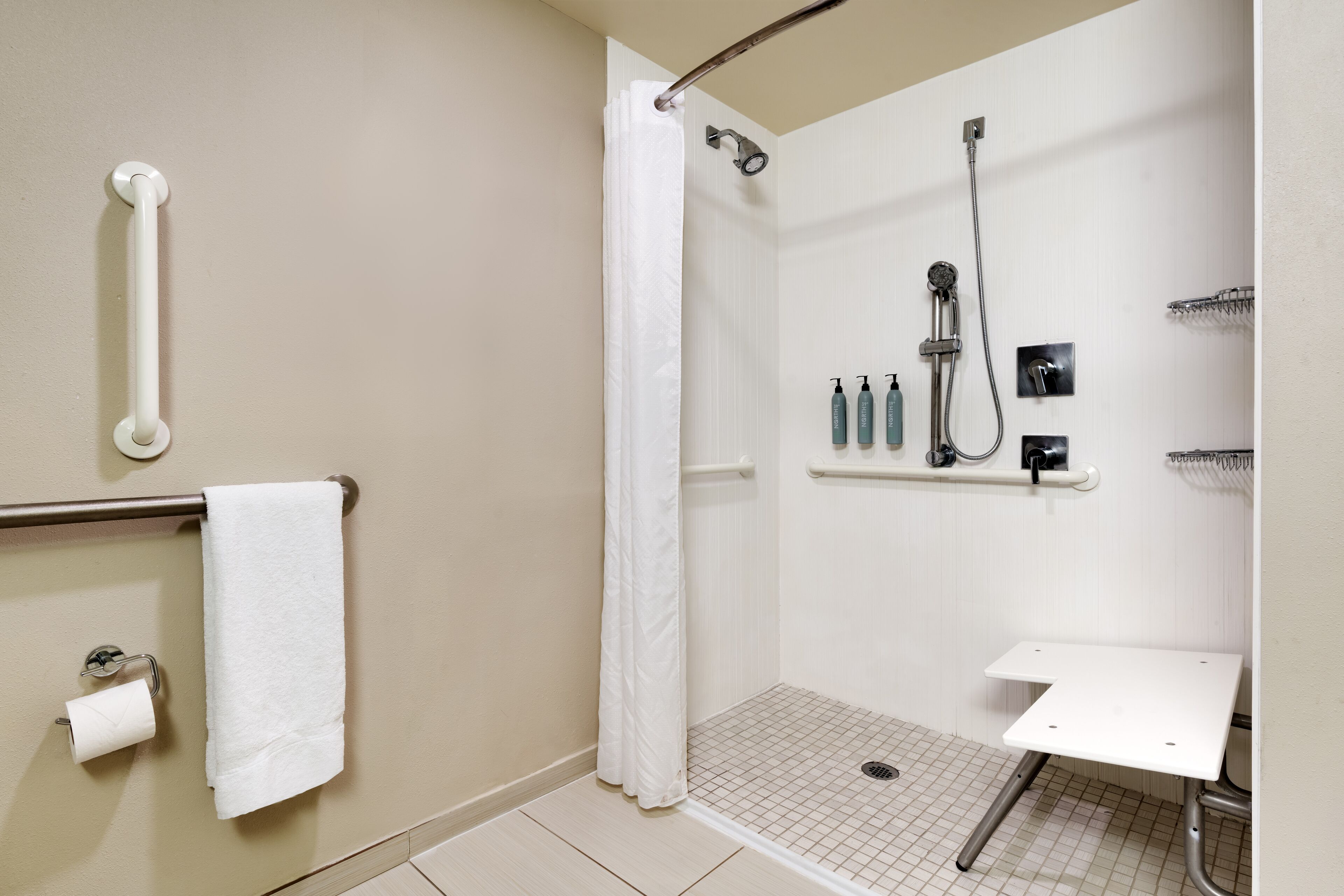 A bathroom with a shower, grab bar, toilet paper, and a shelf with toiletries.