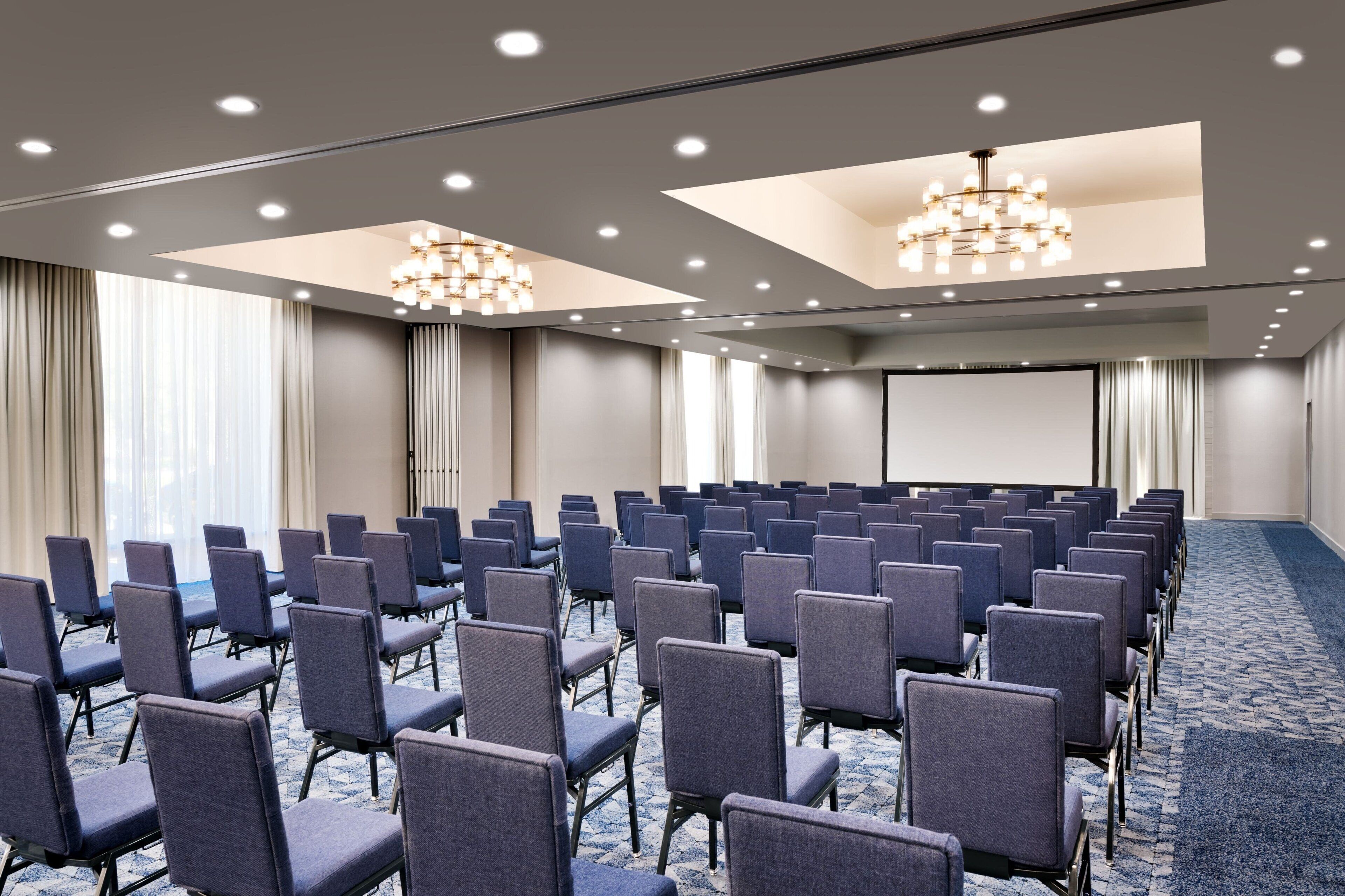 Un salón de conferencias con filas de sillas, una pantalla de proyector y una gran lámpara de araña.
