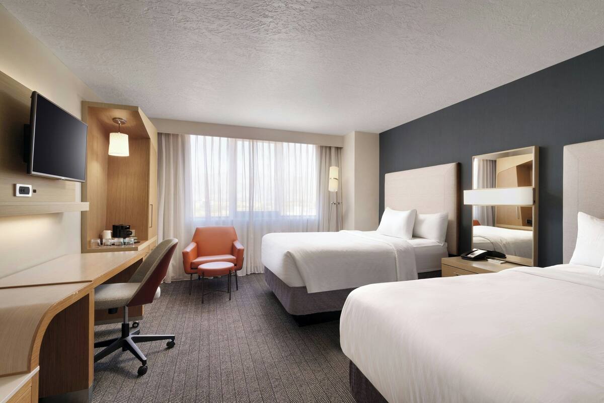 A hotel room with a desk, two beds, a TV, and a large window with curtains.