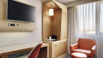 Habitación de hotel con televisor de pantalla plana en la pared, un escritorio de madera con silla, un pequeño otoman, un sillón con reposabrazos y un mueble con lámpara.