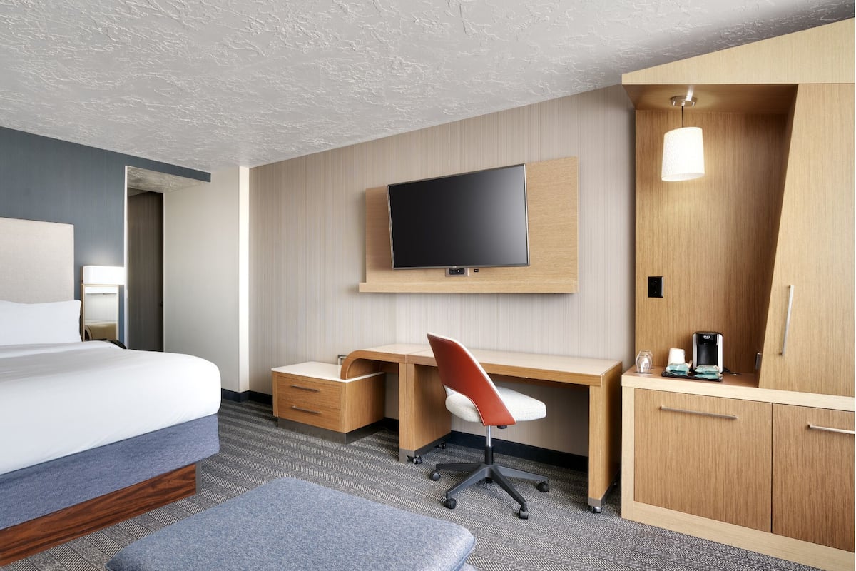 A hotel room with a bed, a desk with a chair, a television, a wardrobe, and a small table with a lamp.