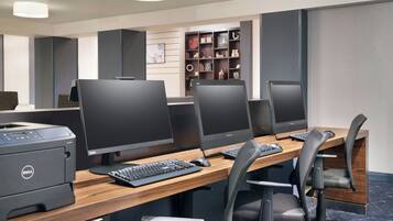 A modern office space with three desks, each equipped with a computer monitor, keyboard, and mouse. There is a Dell printer on the left side of the desks. The room features a wooden desk, black office chairs, and a bookshelf with various items on the wall behind the desks.