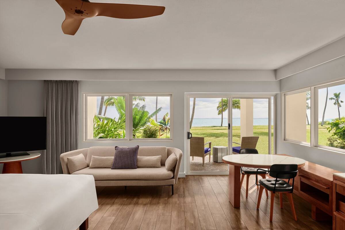 A modern hotel room with a sofa, TV, dining table, and chairs. Large windows offer a view of the beach and palm trees.