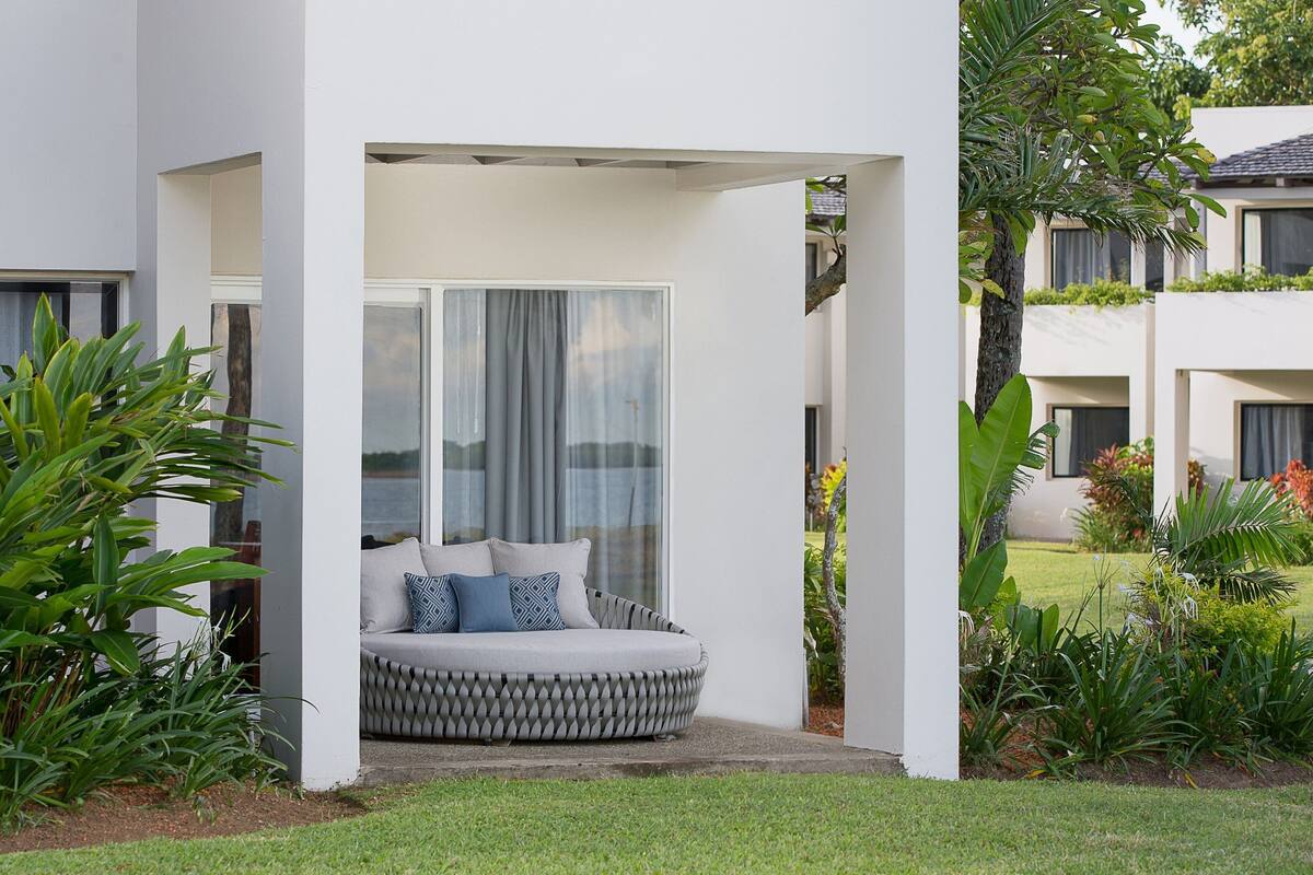 A modern outdoor seating area with a cushioned bench and a glass door, surrounded by lush greenery.