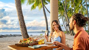 A wooden table with a meal and drinks on a beach with palm trees and the ocean in the background. - Sheraton Fiji Golf & Beach Resort (Nadi)