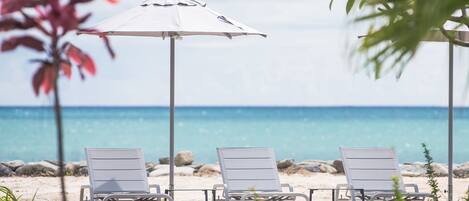 A poolside area with three lounge chairs and an umbrella, overlooking a beach with the ocean in the background.