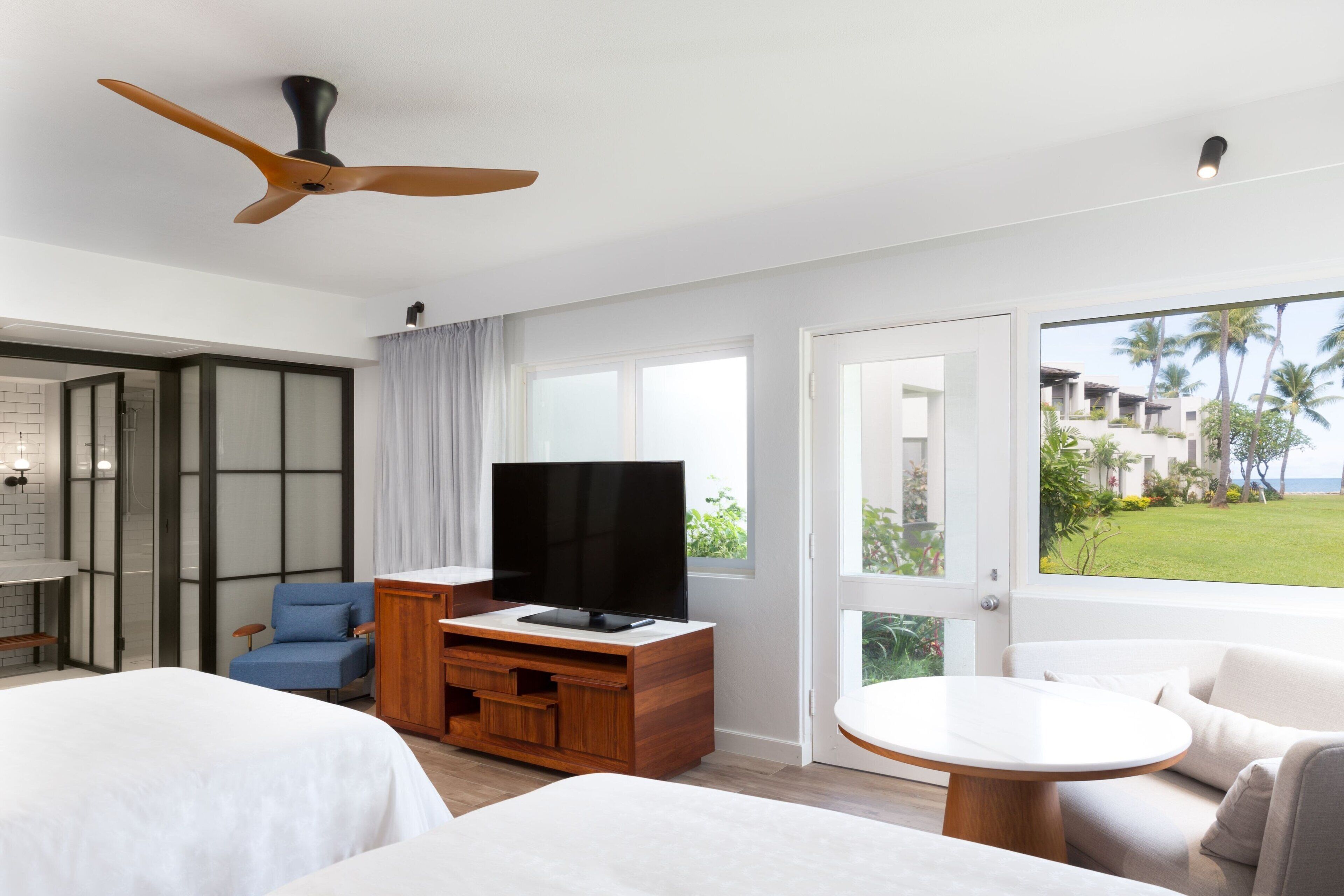 A modern hotel room with a large bed, a TV on a wooden stand, a seating area with a round table, and a balcony with a view of palm trees.