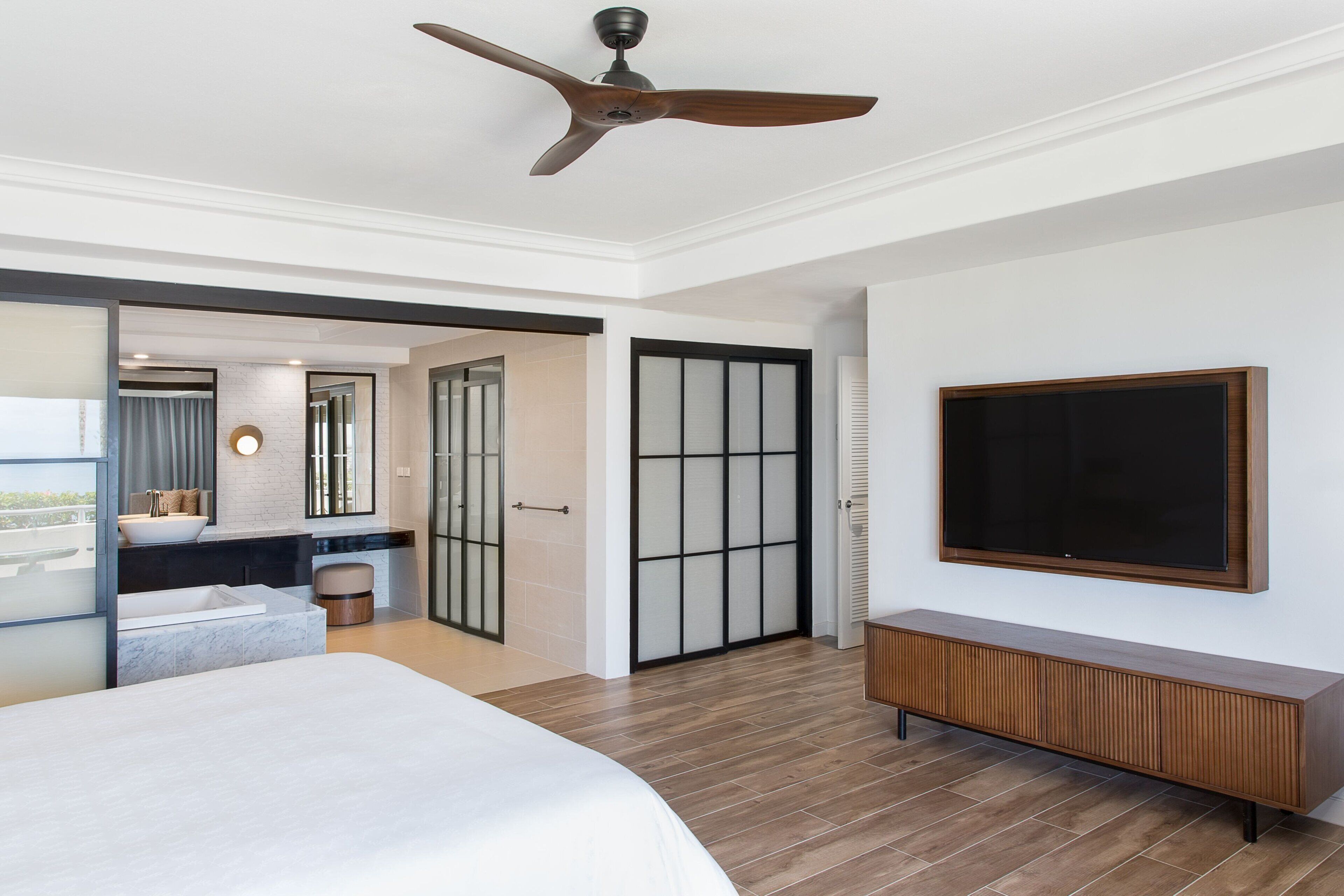 A modern hotel room with a large bed, a ceiling fan, a TV mounted on the wall, and a sliding door leading to a bathroom.