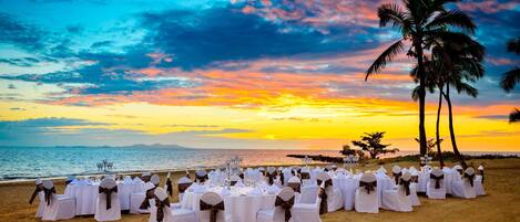 A beach wedding setup with round tables and chairs, palm trees, and a sunset view.