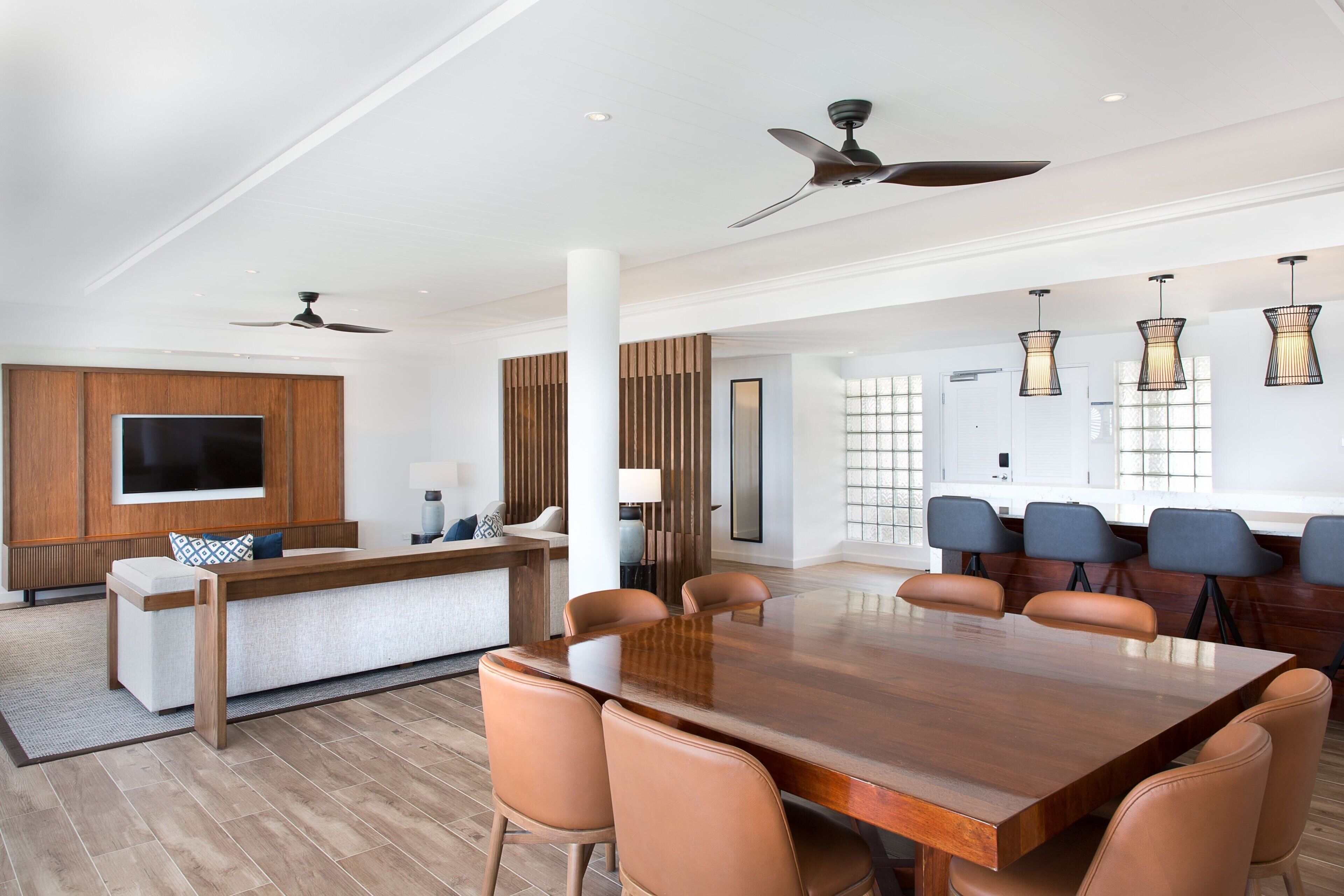 A modern dining area with a large wooden table and leather chairs, a TV mounted on a wooden panel, and ceiling fans.