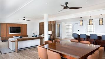 A modern dining area with a large wooden table and leather chairs, a TV mounted on a wooden panel, and ceiling fans.