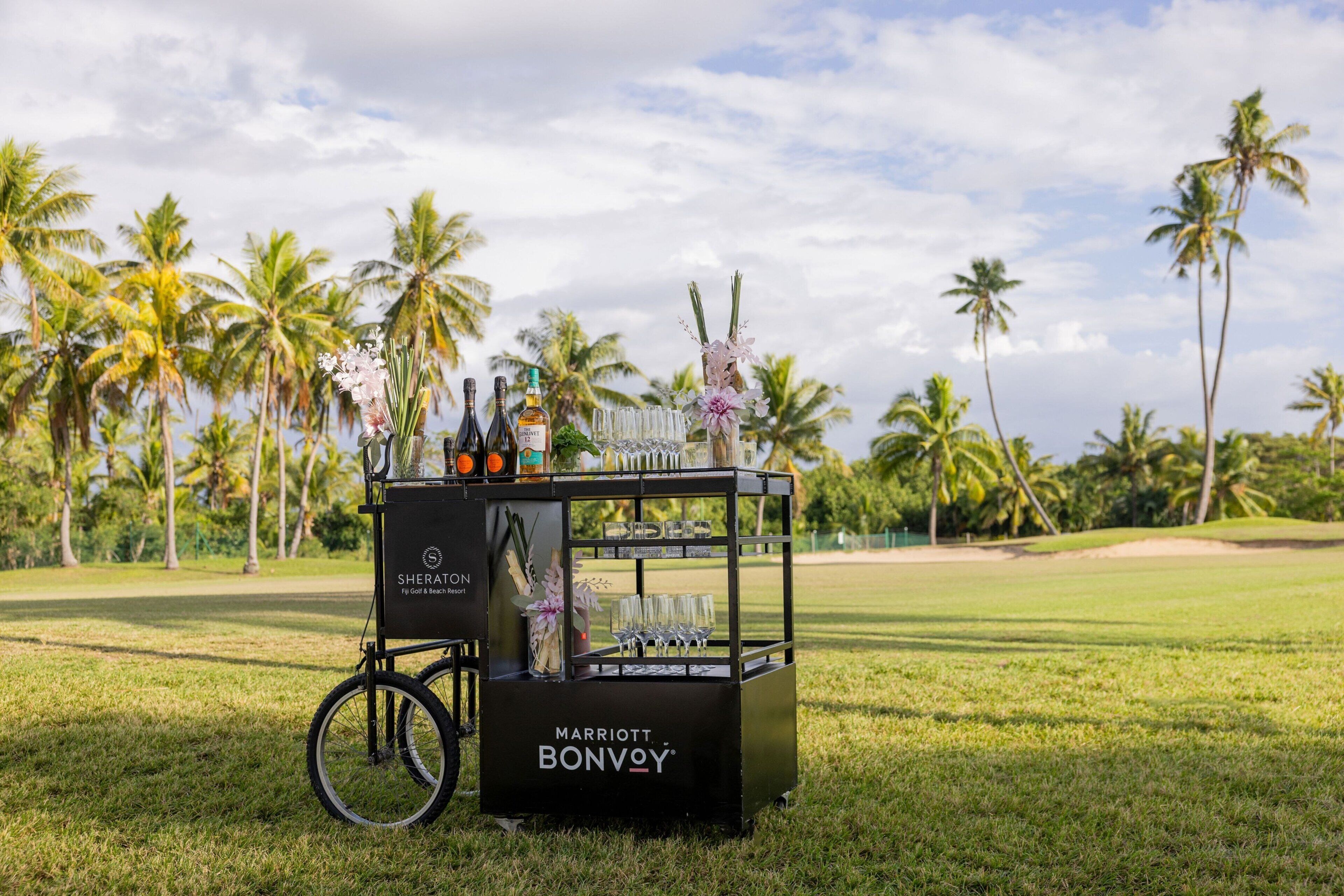 Un chariot portant une pancarte indiquant « Sheraton Fly Golf & Beach Resort » et « Marriott Bonvoy », sur un terrain de golf avec des palmiers en arrière-plan.