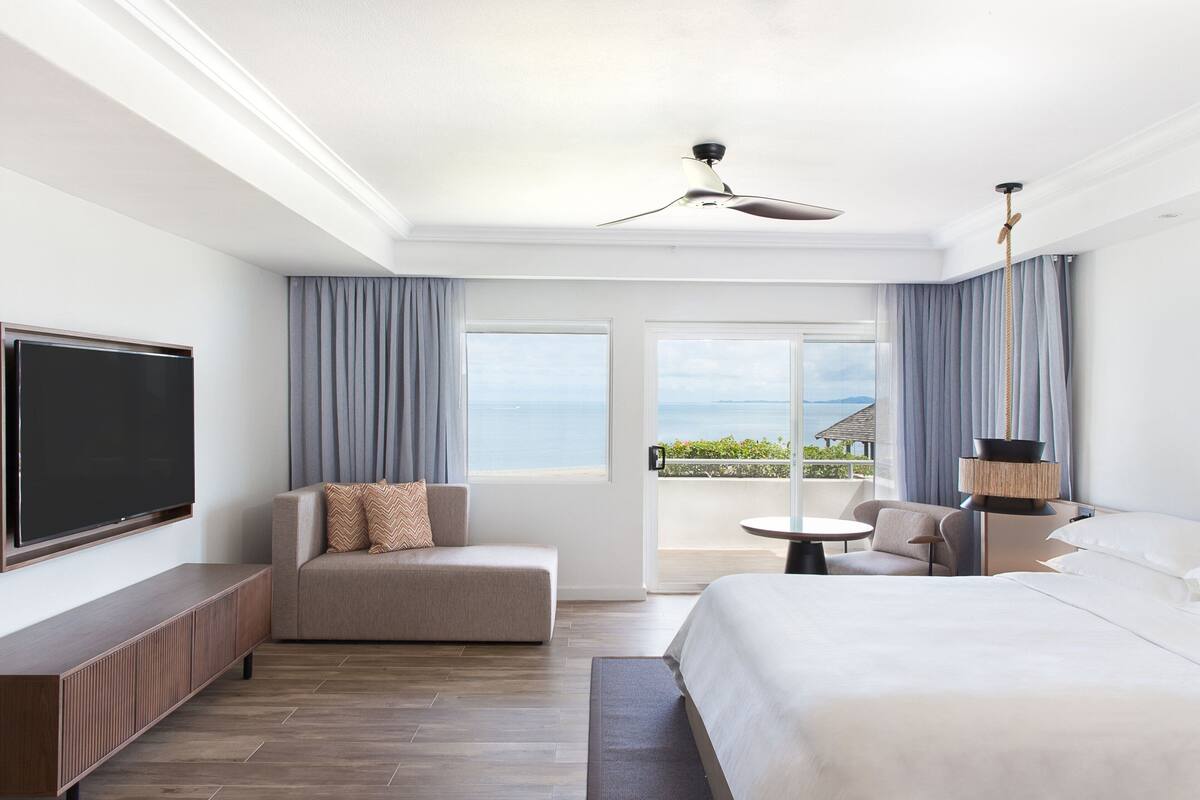 A hotel room with a bed, a sofa, a TV, and a balcony with a view of the sea.