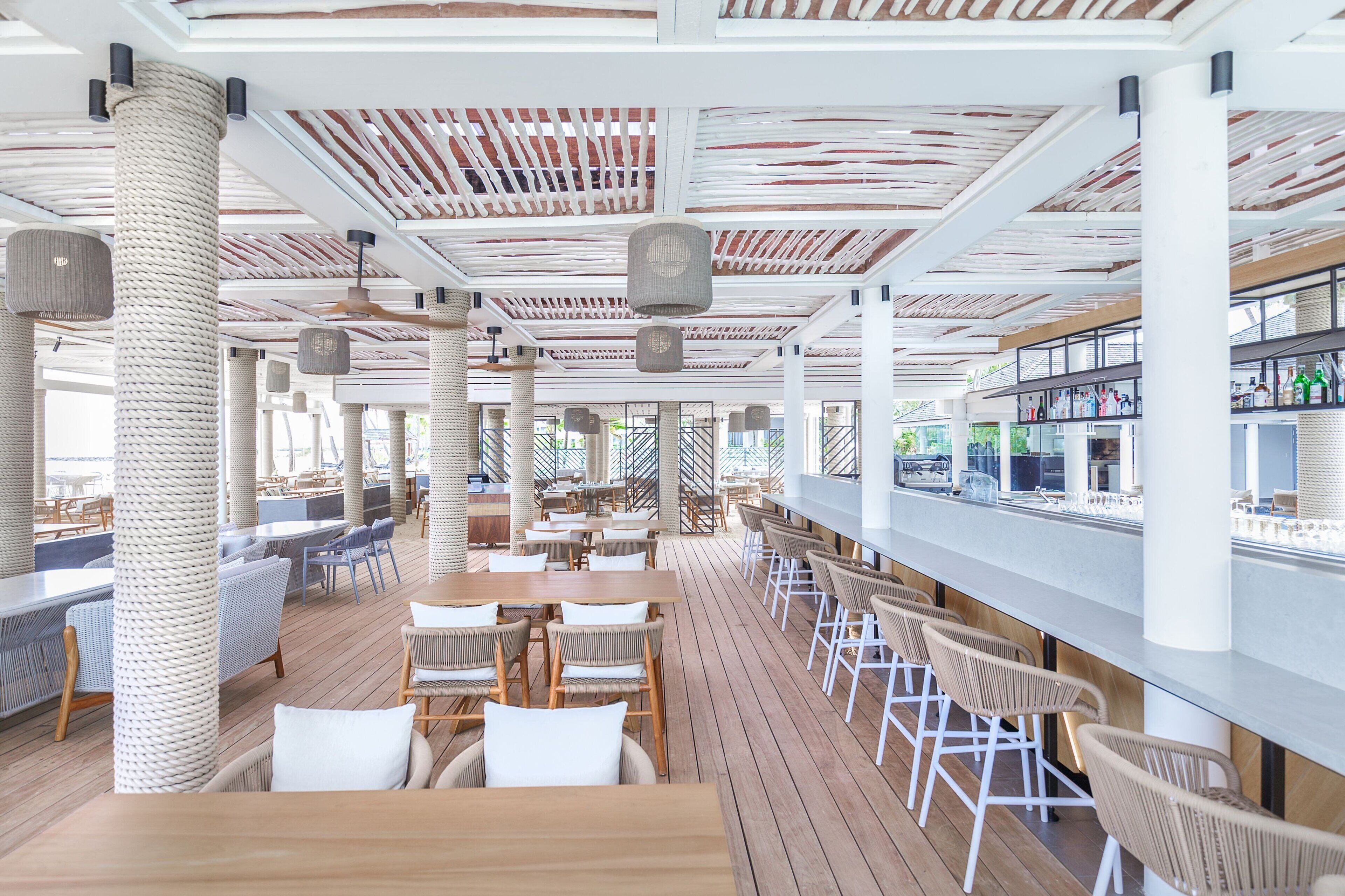 Un amplio espacio de comedor al aire libre con mesas de madera y sillas de mimbre, una barra con una larga encimera y un techo con diseño de listones de madera.