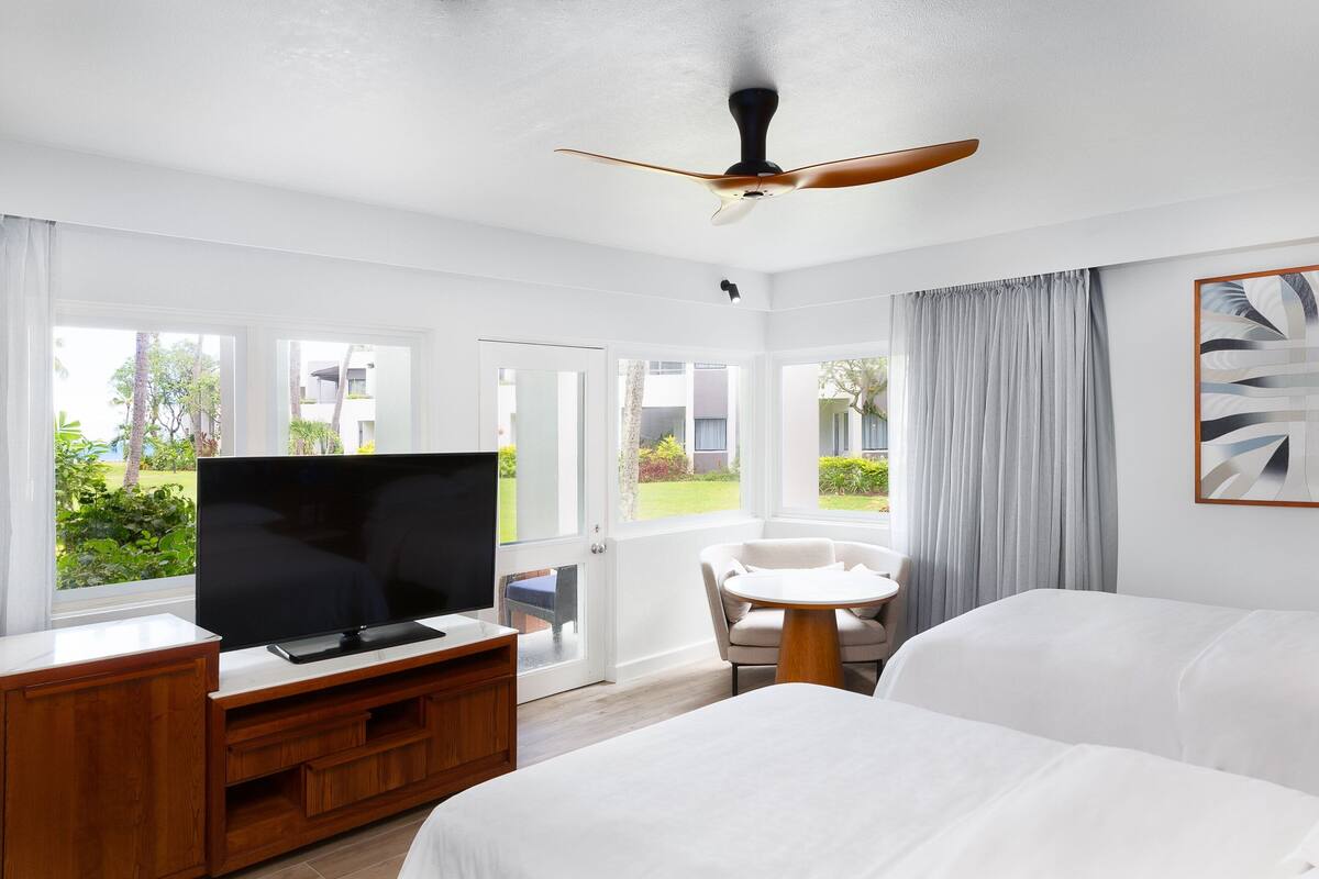 A hotel room with a bed, a TV on a wooden stand, a ceiling fan, and a view of the outdoors through large windows.