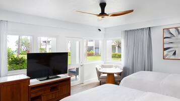 A hotel room with a bed, a TV on a wooden stand, a ceiling fan, and a view of the outdoors through large windows.