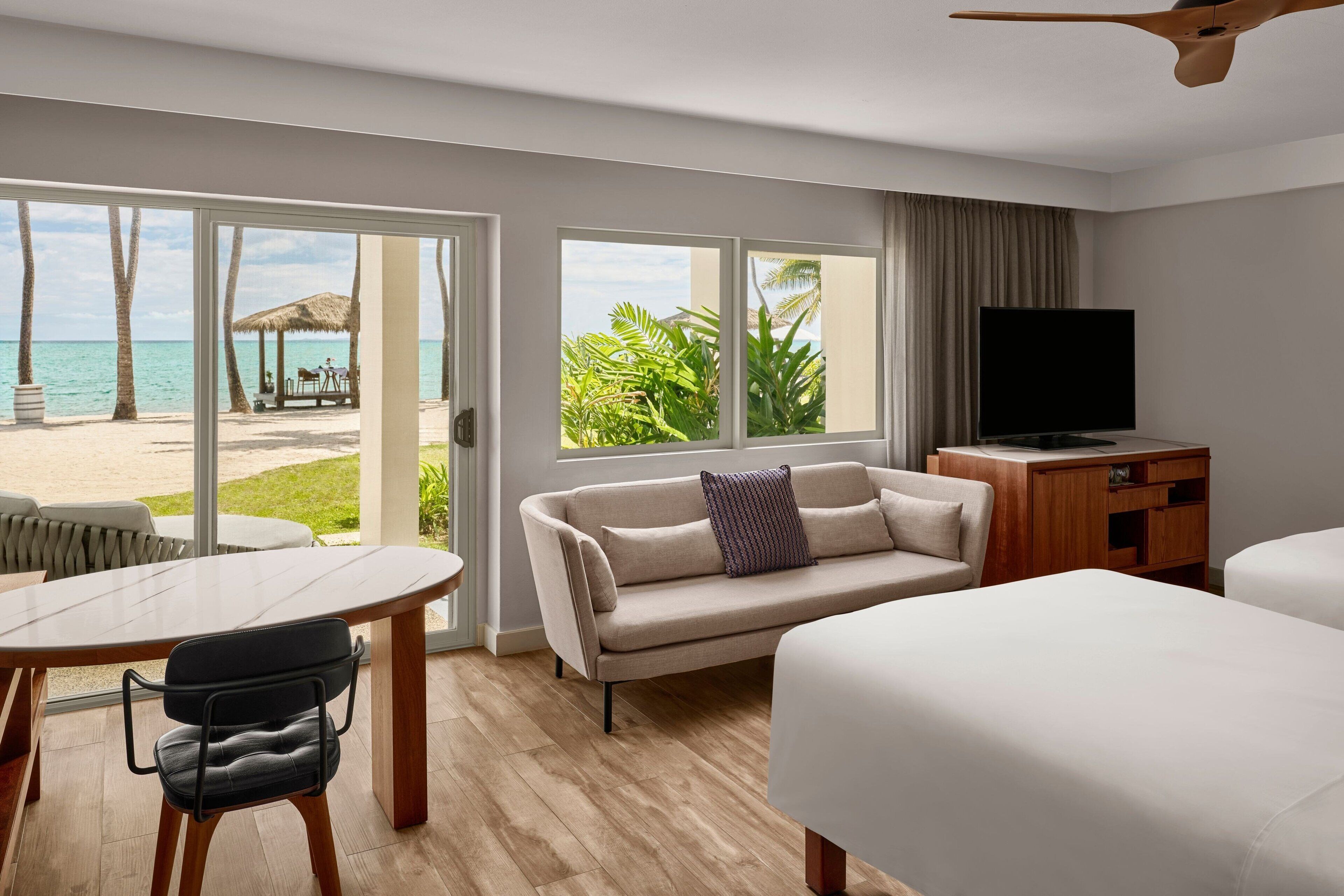 A hotel room with a large window offering a view of the beach and ocean, a sofa, a TV, and a dining area with a table and chairs.