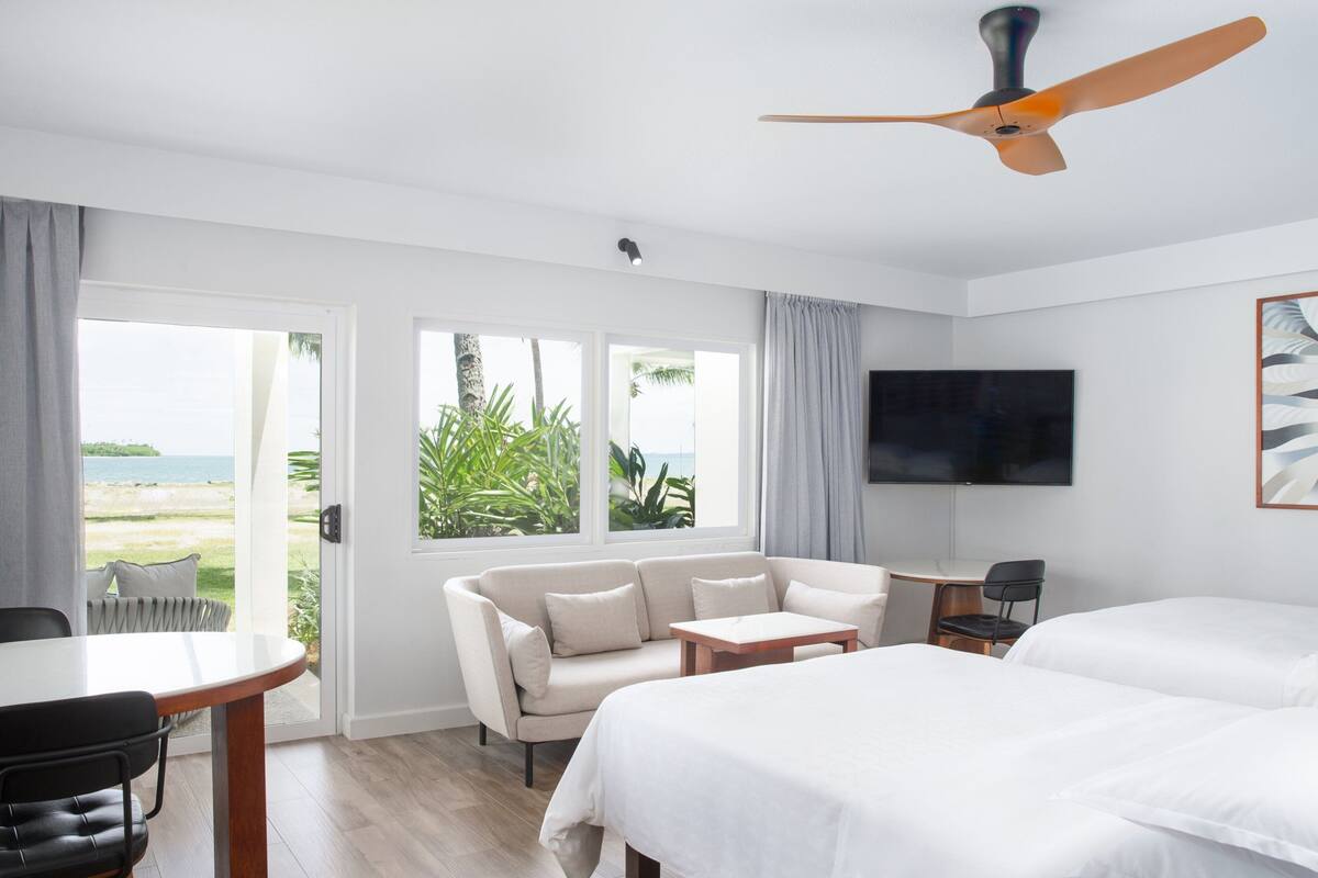 A hotel room with a bed, a sofa, a TV, a ceiling fan, and a view of the beach.