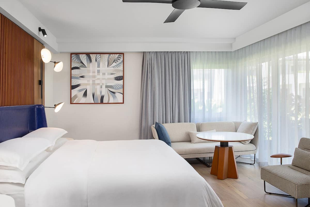 A modern hotel room with a large bed, a sofa, a small table, and a ceiling fan.