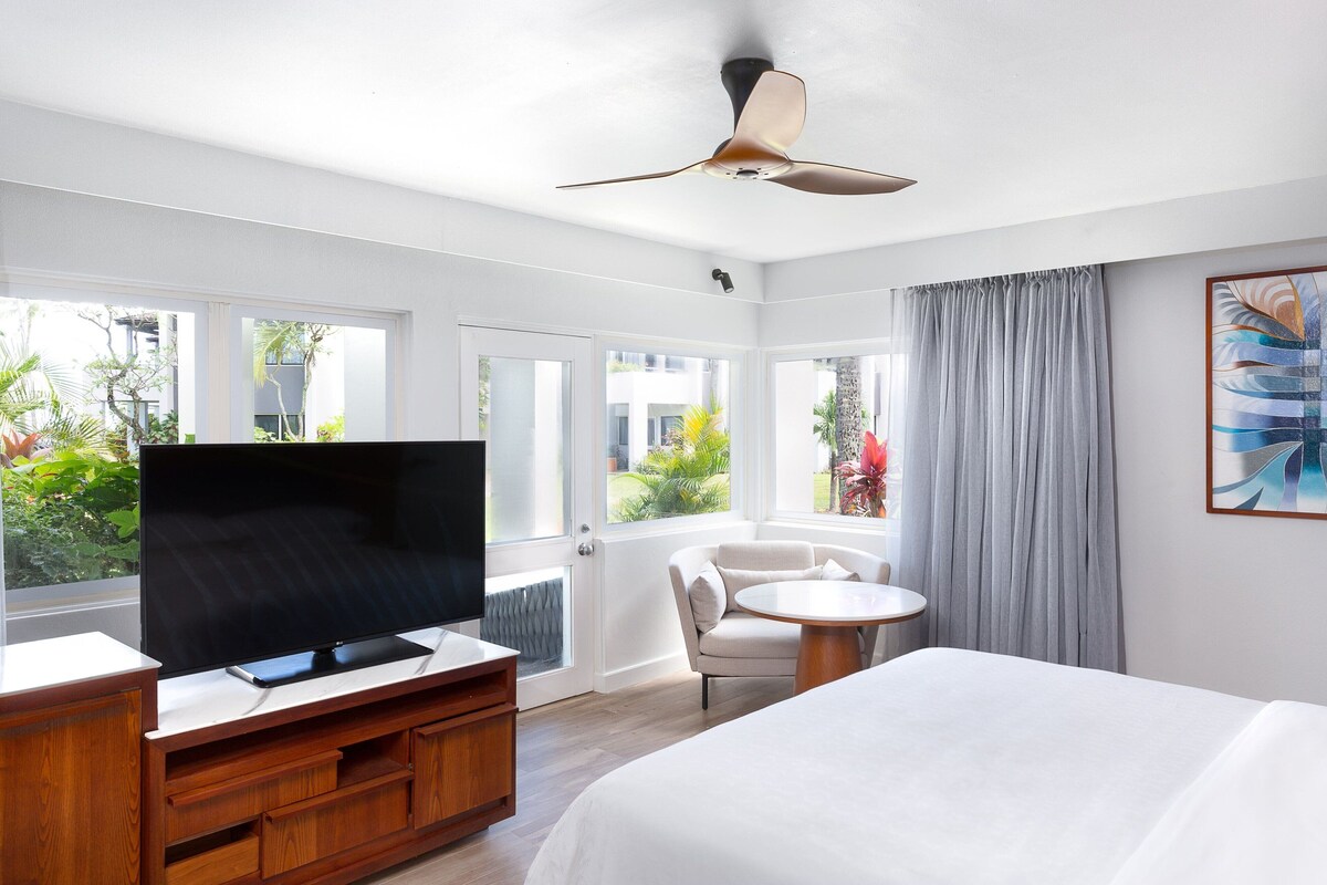 A hotel room with a bed, a TV on a wooden stand, a chair, a small table, and a ceiling fan.