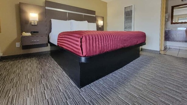 A hotel room with a bed, a nightstand, a wall-mounted lamp, and a bathroom visible through an open door. - Americas Best Value Inn Williams Grand Canyon (Williams)
