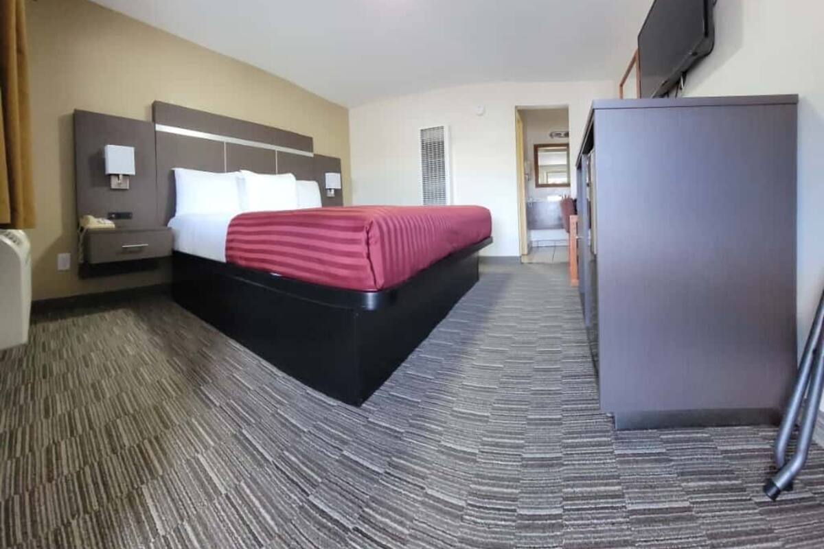 A hotel room with a bed, a desk, a flat-screen TV, a refrigerator, and a carpeted floor.