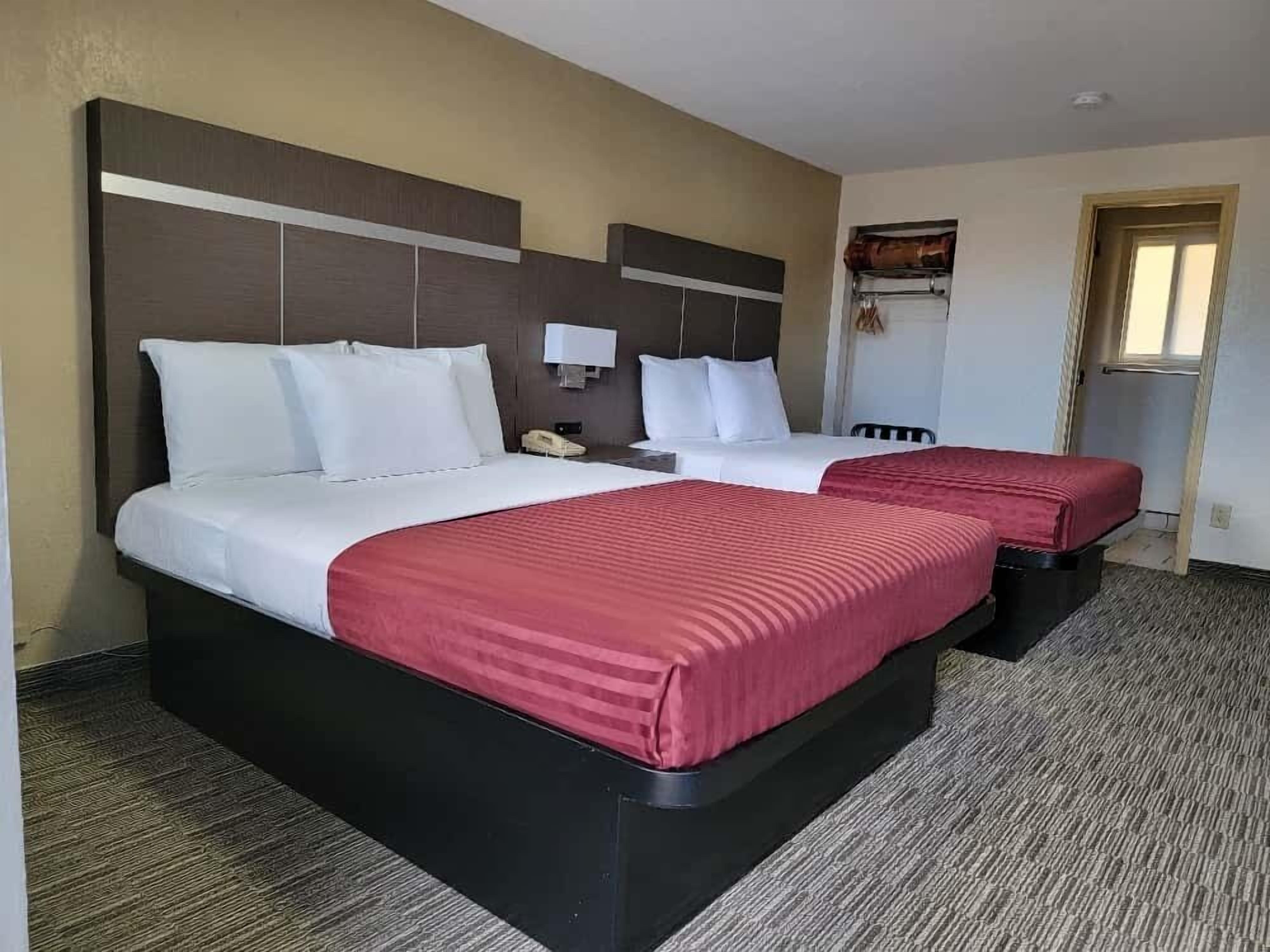 Habitación de hotel con dos camas, cada una con ropa blanca y detalles rojos, una mesita de noche con lámpara y una ventana con vistas.