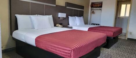 A hotel room with two beds, each with white bedding and red accents, a small bedside table with a lamp, and a window with a view.