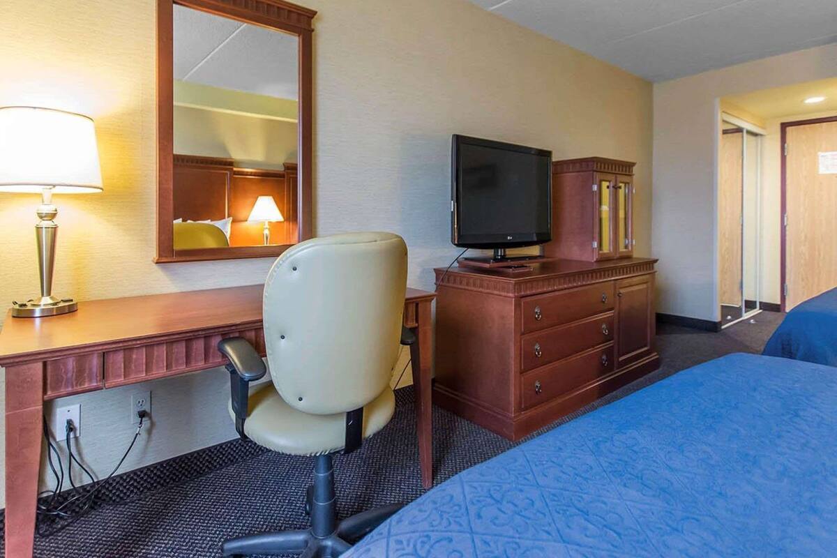 Une chambre d’hôtel avec un lit, un bureau avec une lampe, une chaise, un téléviseur posé sur une commode en bois et un miroir au mur.