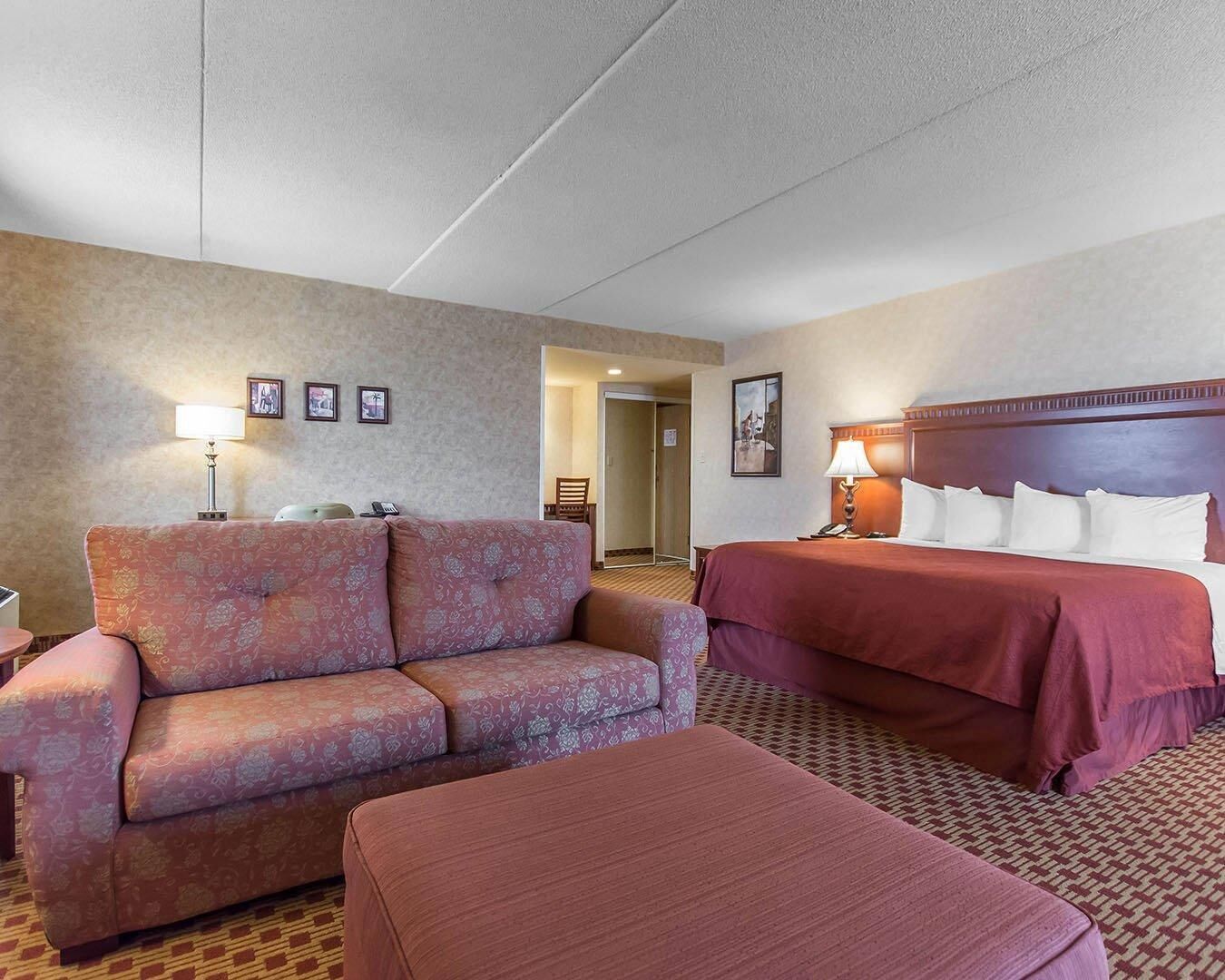 Una habitación de hotel con sofá, cama, reposapiés y escritorio con lámpara.