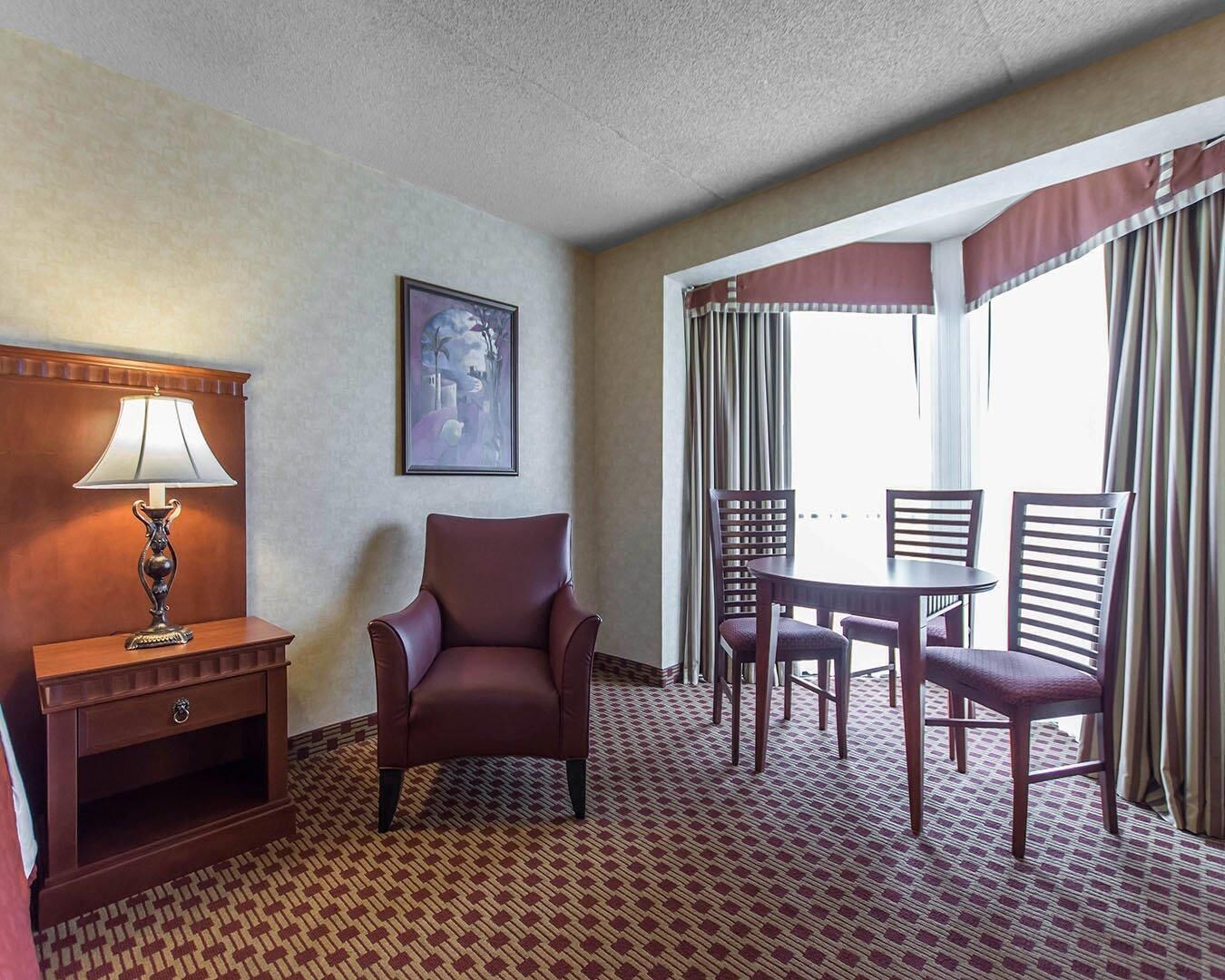 A hotel room with a bed, a desk with a lamp, a chair, a dining table with chairs, and a large window with curtains.