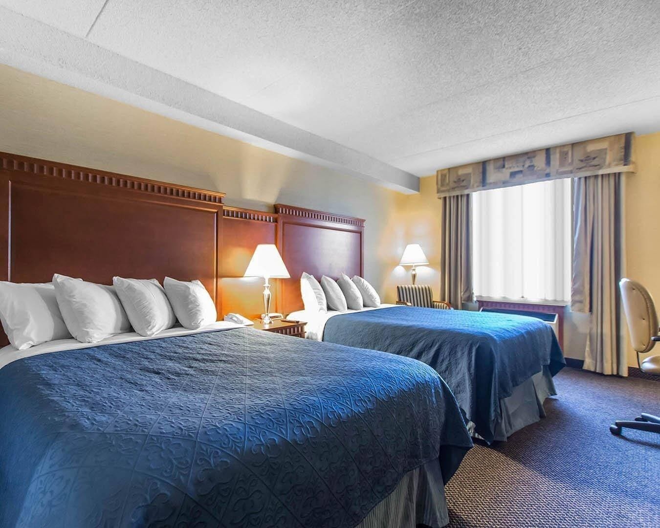 Une chambre d’hôtel avec deux lits, une tête de lit en bois, un bureau avec une lampe, une chaise et une fenêtre avec des rideaux.