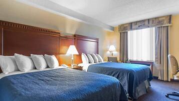 Une chambre d’hôtel avec deux lits, une tête de lit en bois, un bureau avec une lampe, une chaise et une fenêtre avec des rideaux.