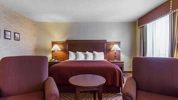 A hotel room with a bed, two bedside tables with lamps, a small round table, two armchairs, and a patterned carpet.