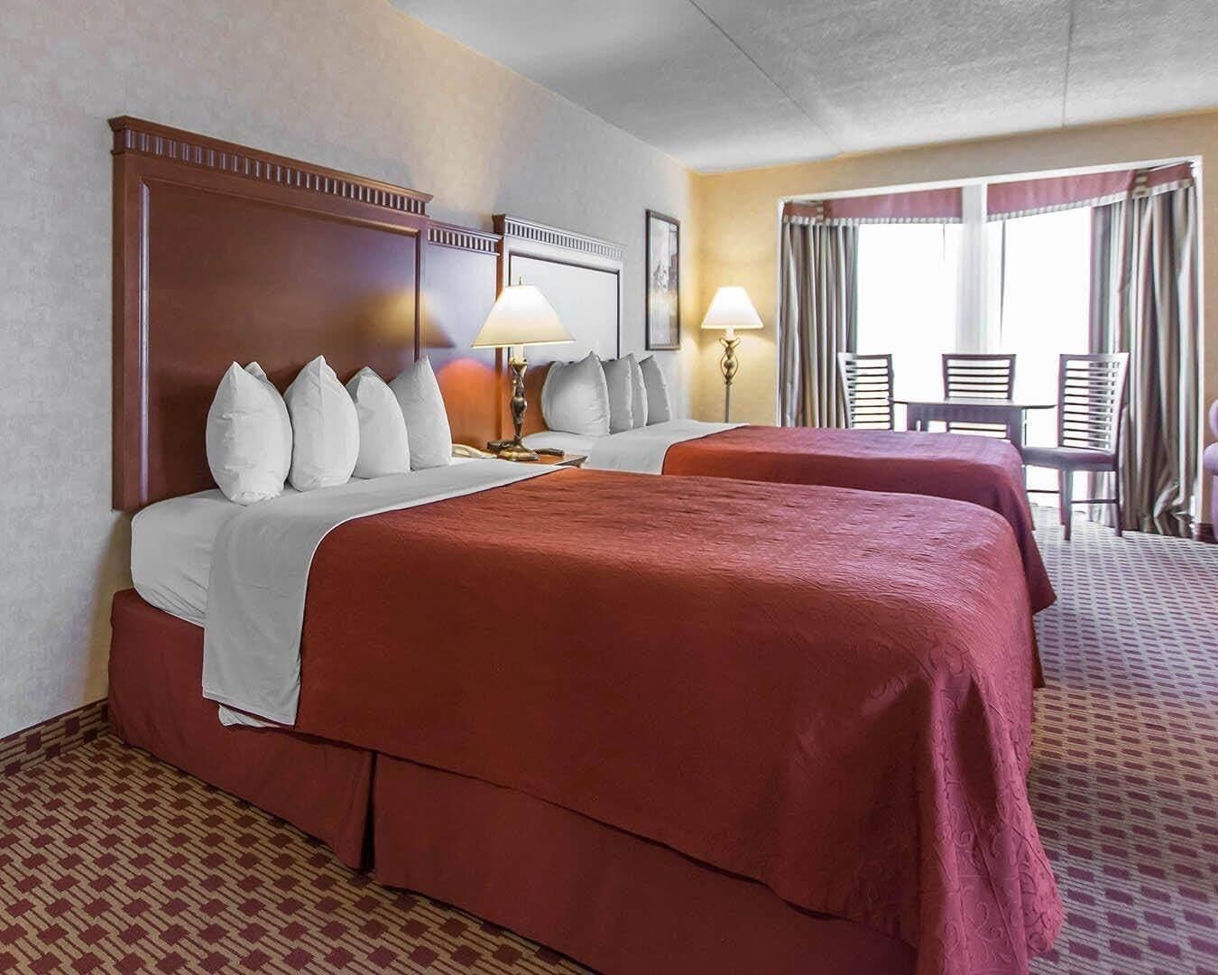 Habitación de hotel con dos camas, una zona de estar con mesa y sillas, y un ventanal con cortinas.